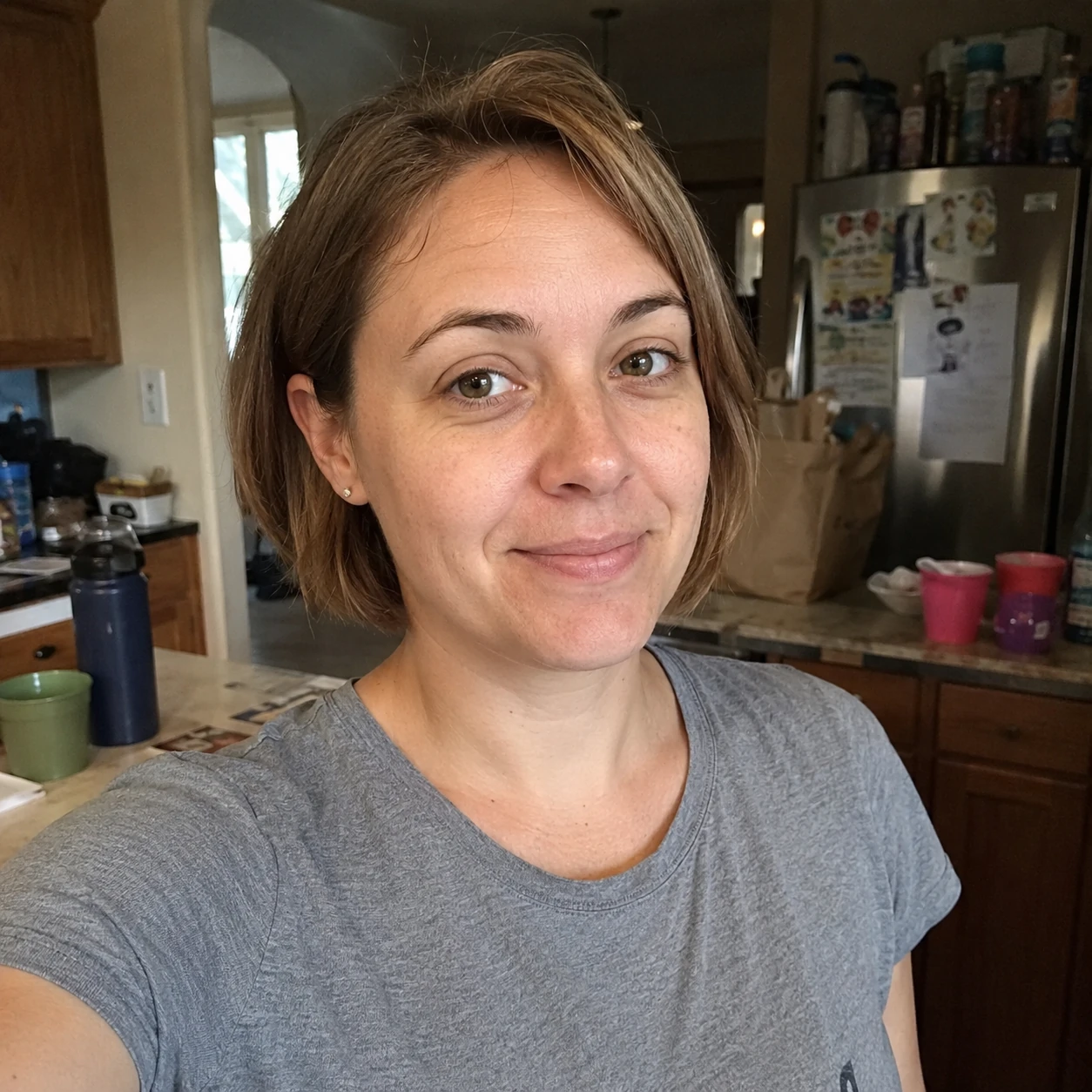 31-year-old woman with short brown hair and a gray T-shirt standing in a slightly cluttered family kitchen.