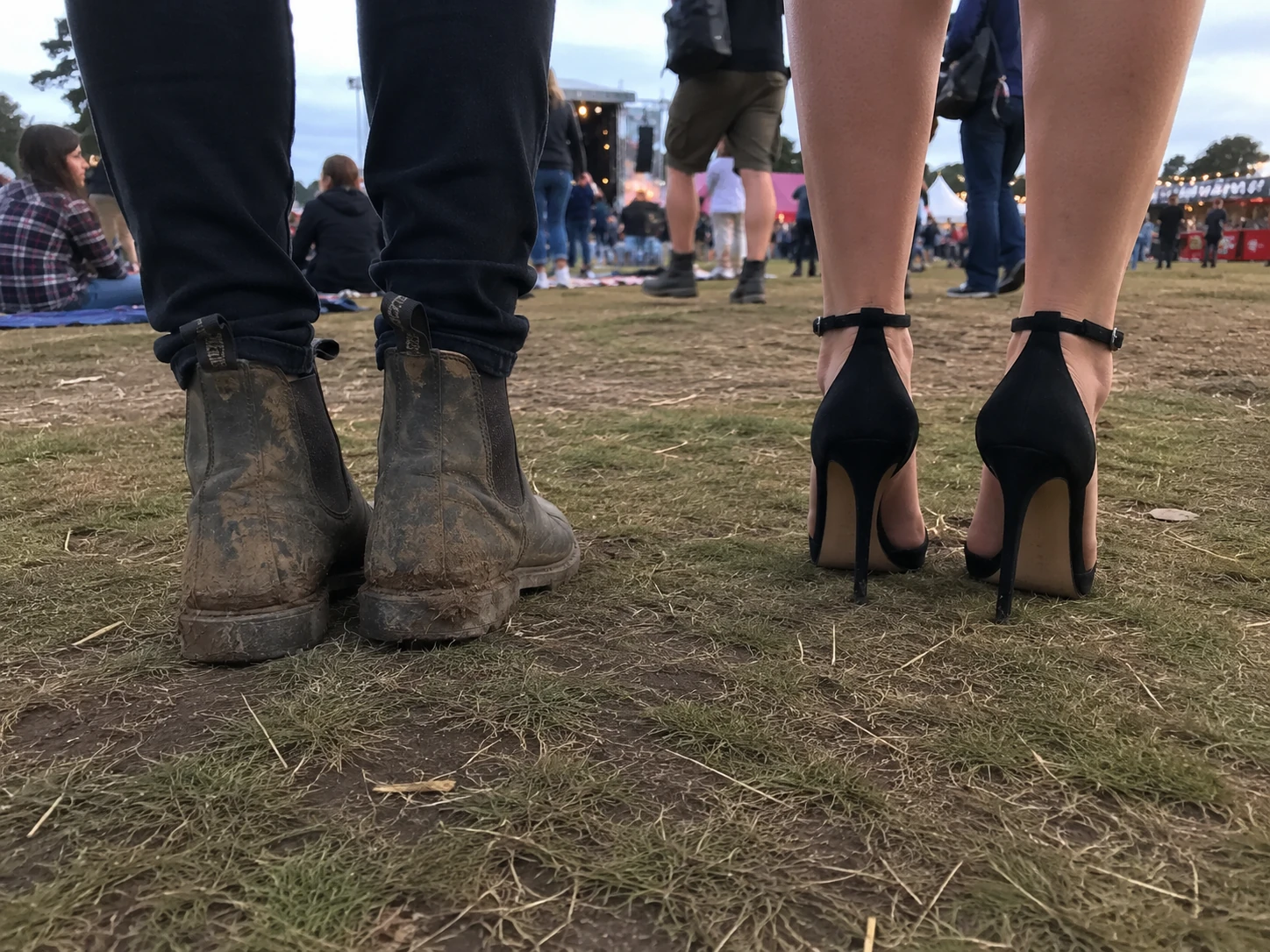 Two women stand on a festival field, one wearing mud-covered practical boots and the other wearing spotless, extremely high black stiletto heels.