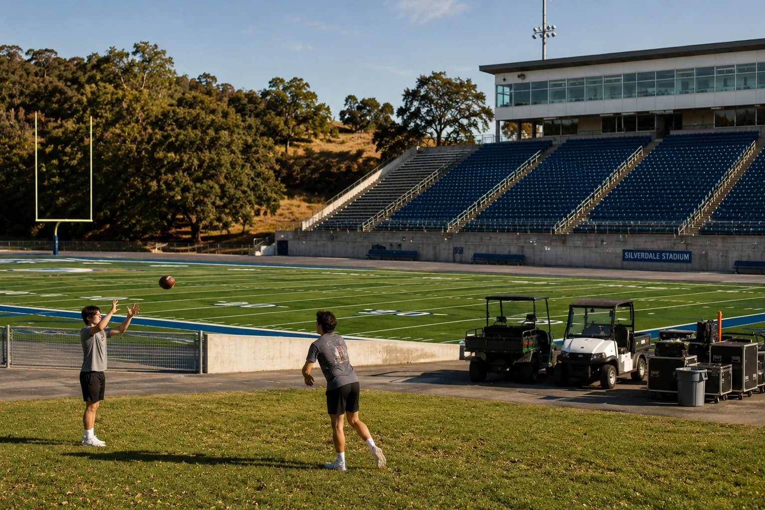 A casual documentary-style photograph of Silverdale Stadium in Northern California in late-August afternoon light, a few staff carts and equipment cases near the sideline, freshly painted football field markings, empty blue-and-green stands, and two college students casually throwing a football badly on a patch of grass outside the stadium. Mature Northern California oaks and dry late-summer vegetation in the background. Ordinary smartphone/news photograph, slightly imperfect framing, no posed players, no readable logos or text.
