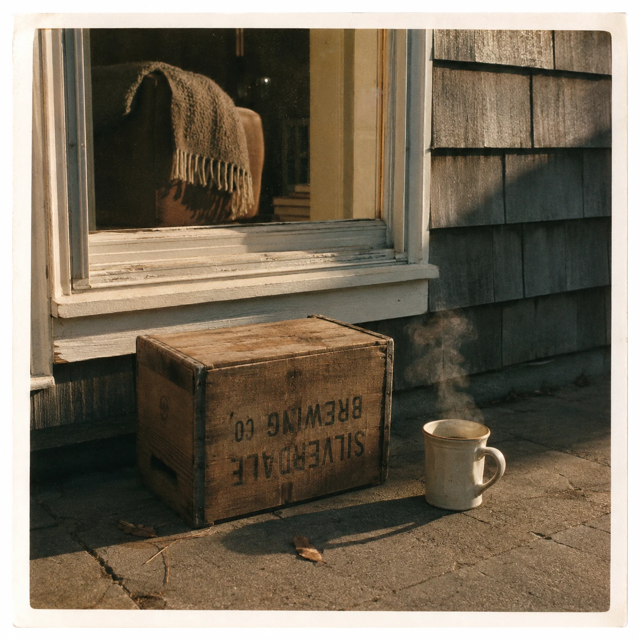 An empty upside-down beer crate beside a cooling mug of coffee outside a quiet suburban home in soft afternoon light, suggesting its usual occupant is temporarily indoors.